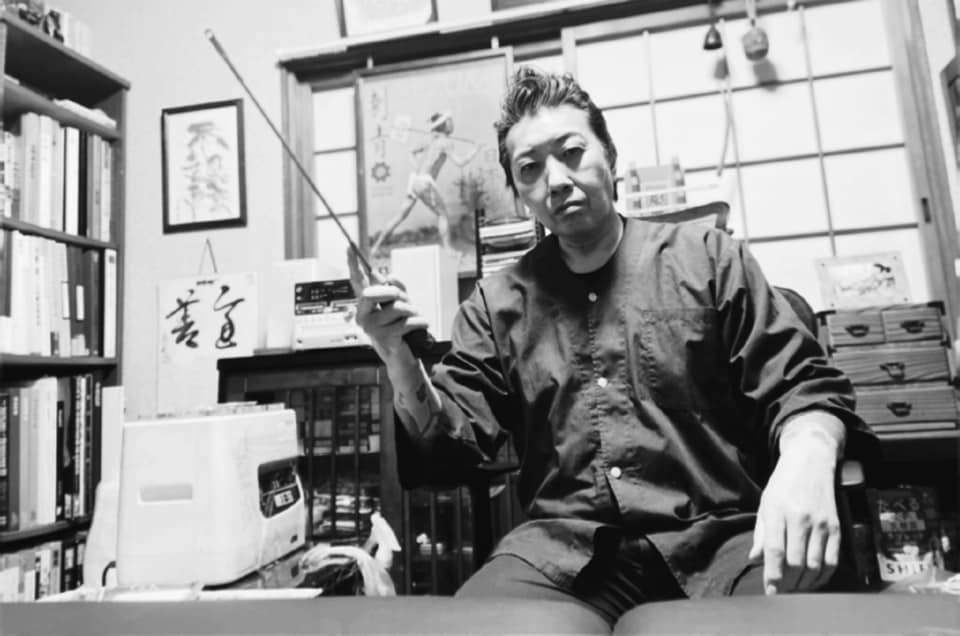 Japanese skin carver Horikoi holding up his Tebori tool. A certain amount of discipline is shown in the picture. In the background you can see pictures of the studio.artefacts of old times are hanging.scrolls from friends and a cabinet full of tools to work with.