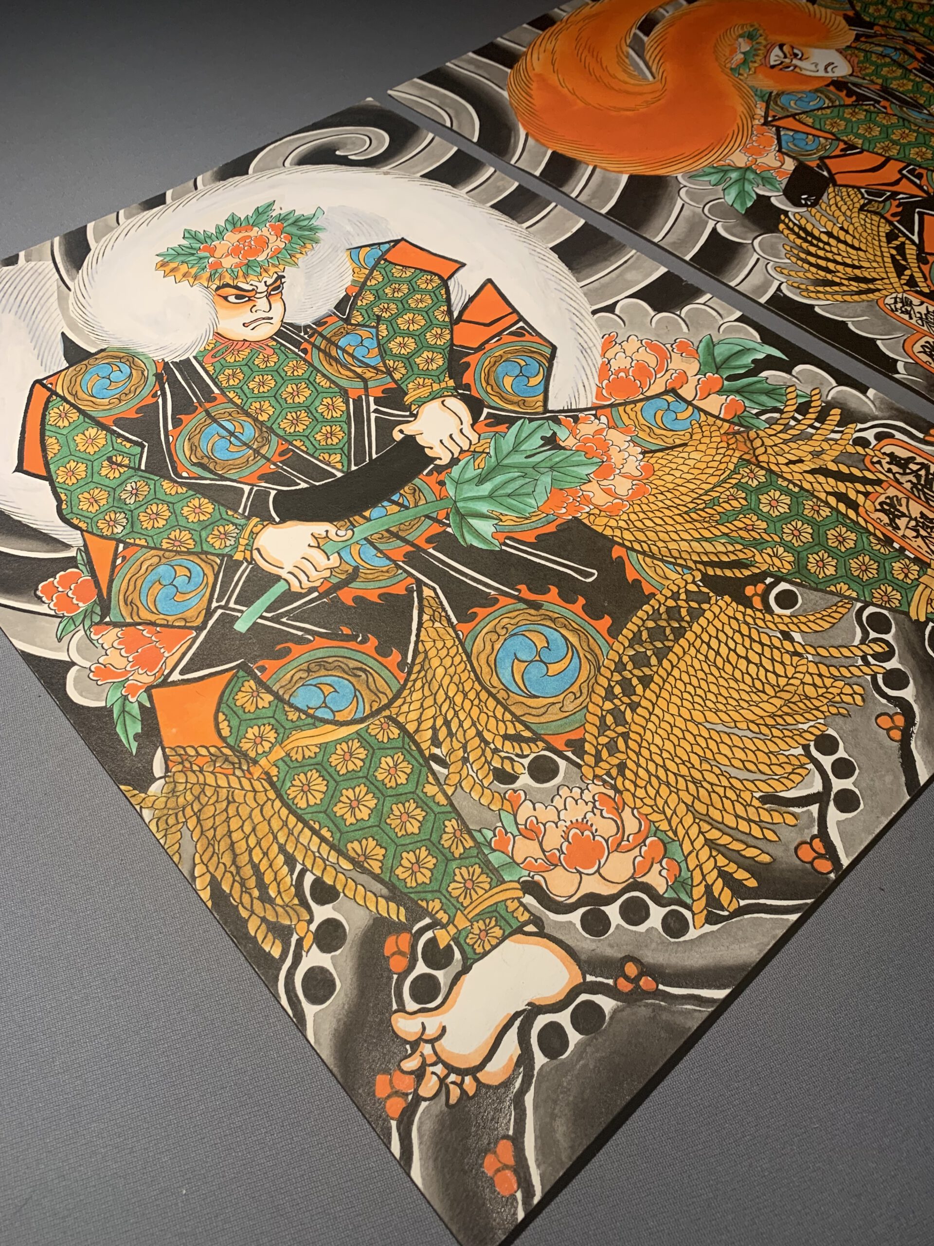 Two Paintings of Kabuki Actors on a gray table. The actors representing Lion dancers from Japanese theater are presented with beautiful garment Design wearing Wings made of Jak ox hair. One has a white wig and the other a red wig. 