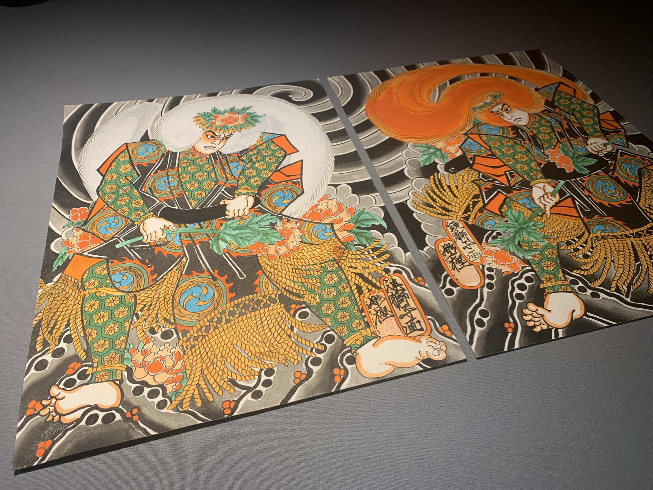 Two Paintings of Kabuki Actors on a gray table. The actors representing Lion dancers from Japanese theater are presented with beautiful garment Design wearing Wings made of Jak ox hair. One has a white wig and the other a red wig. 
