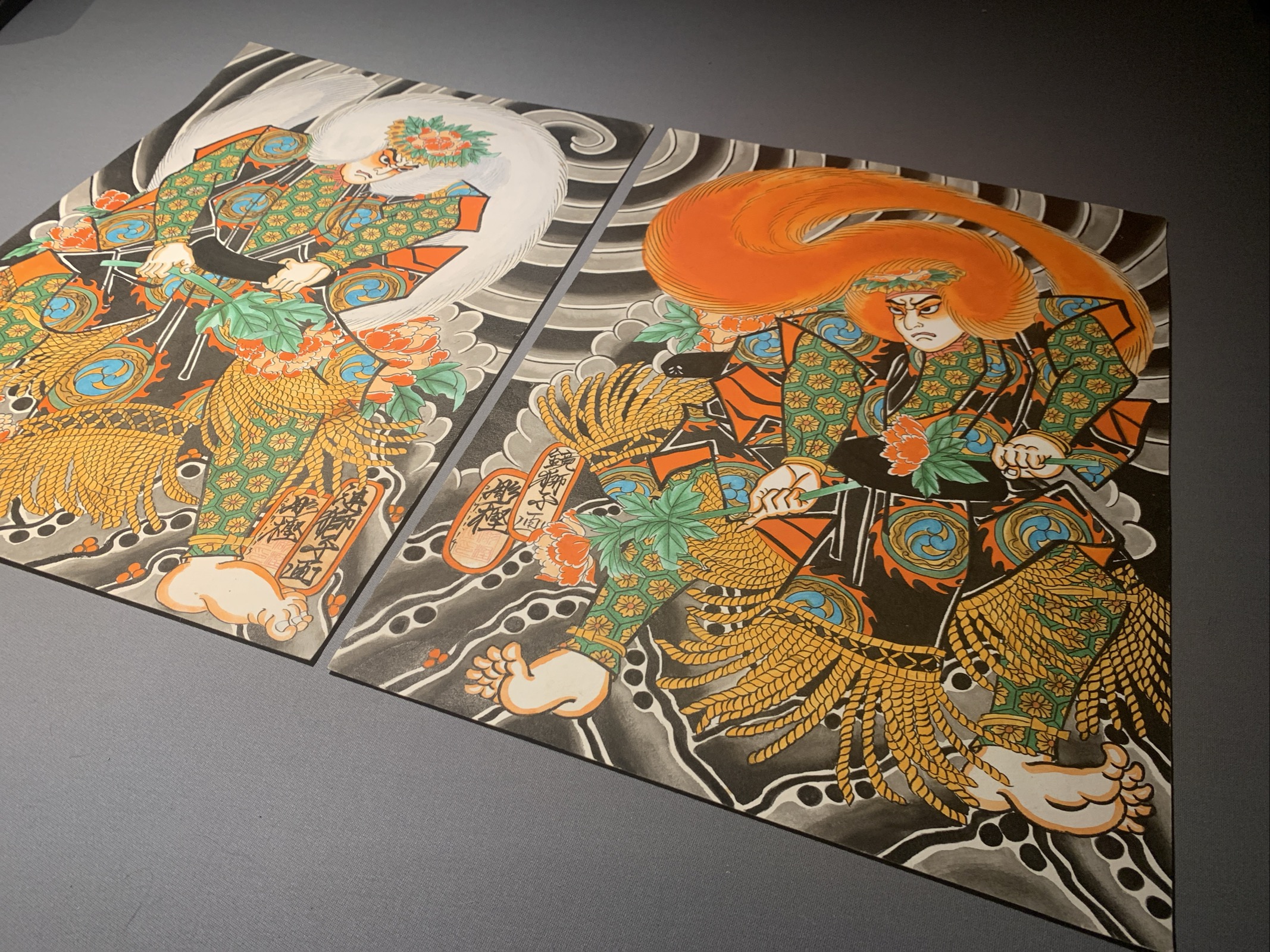 Two Paintings of Kabuki Actors on a gray table. The actors representing Lion dancers from Japanese theater are presented with beautiful garment Design wearing Wings made of Jak ox hair. One has a white wig and the other a red wig. 