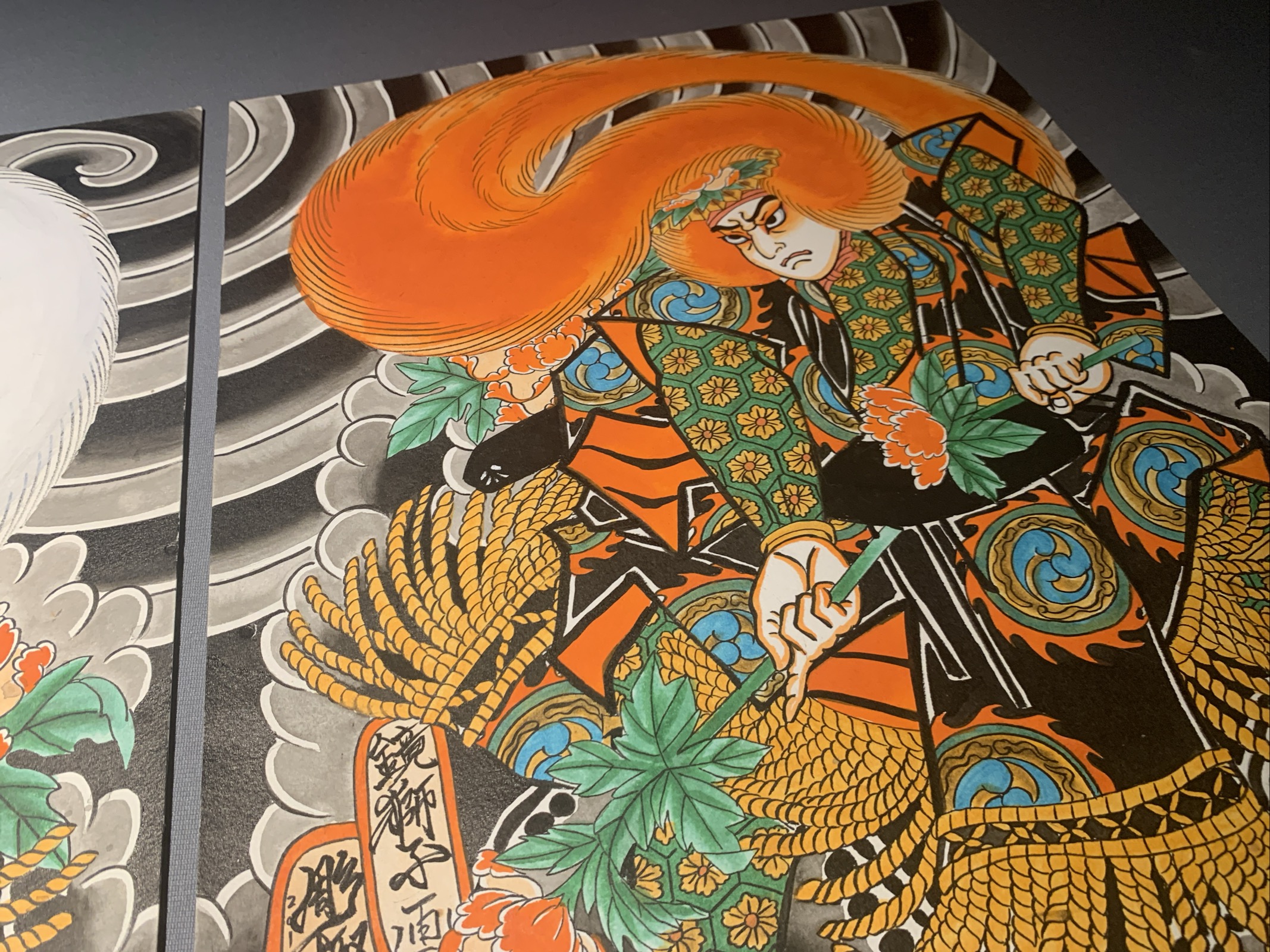 Two Paintings of Kabuki Actors on a gray table. The actors representing Lion dancers from Japanese theater are presented with beautiful garment Design wearing Wings made of Jak ox hair. One has a white wig and the other a red wig. 