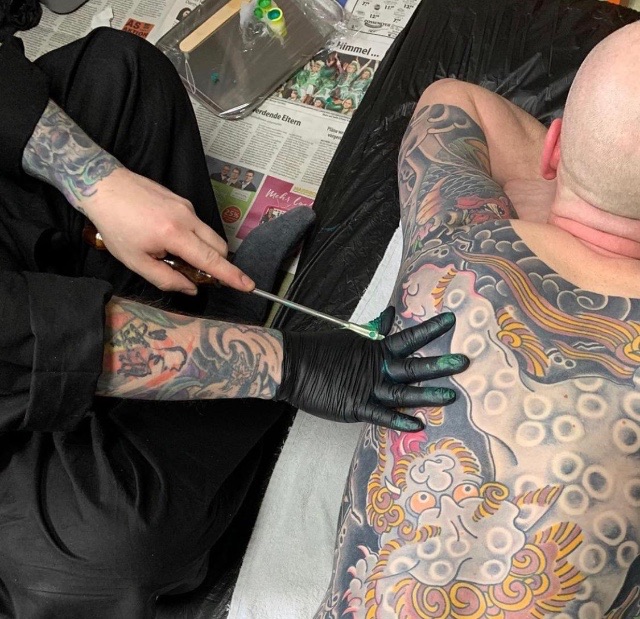 Man with black Gloves inserting ink into another mans body. Shisei the art of tattoo in Japan. A chinese lion can be seen al across the back..