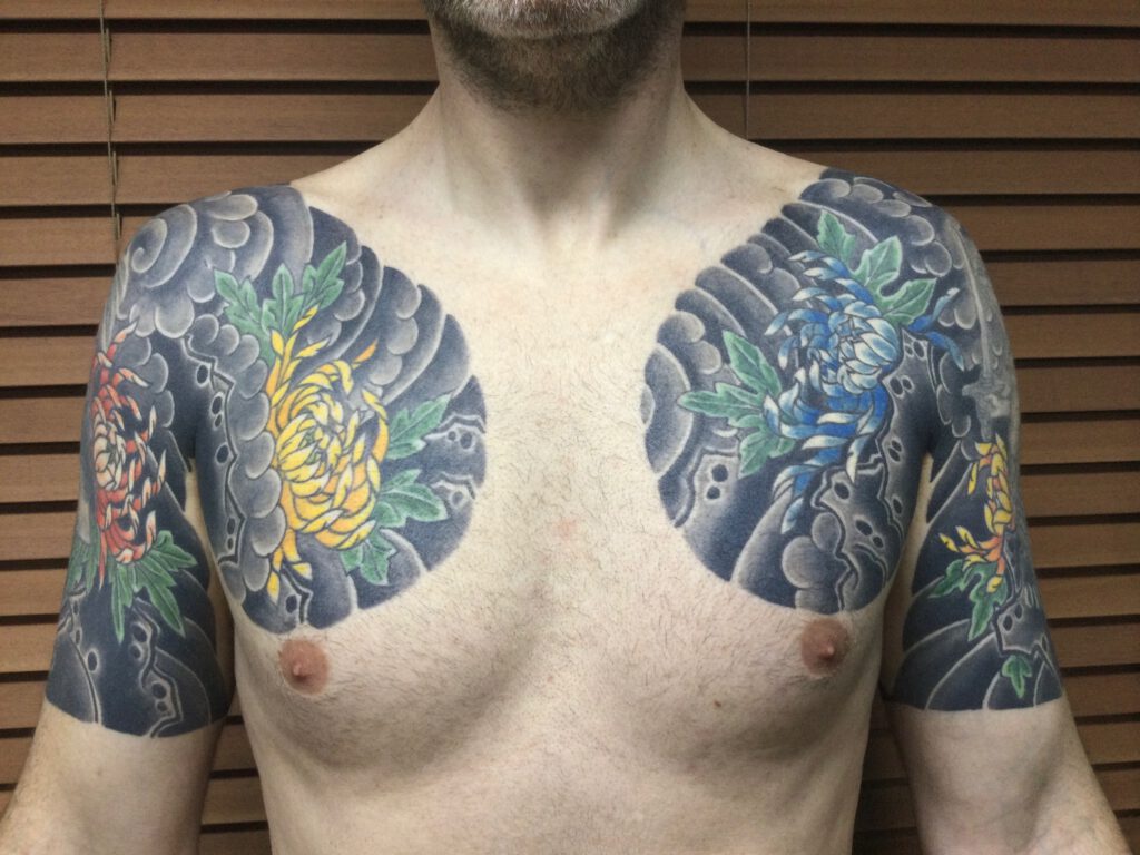 Mans chest and arms both tattooed with Chrysanthemums and Wind.