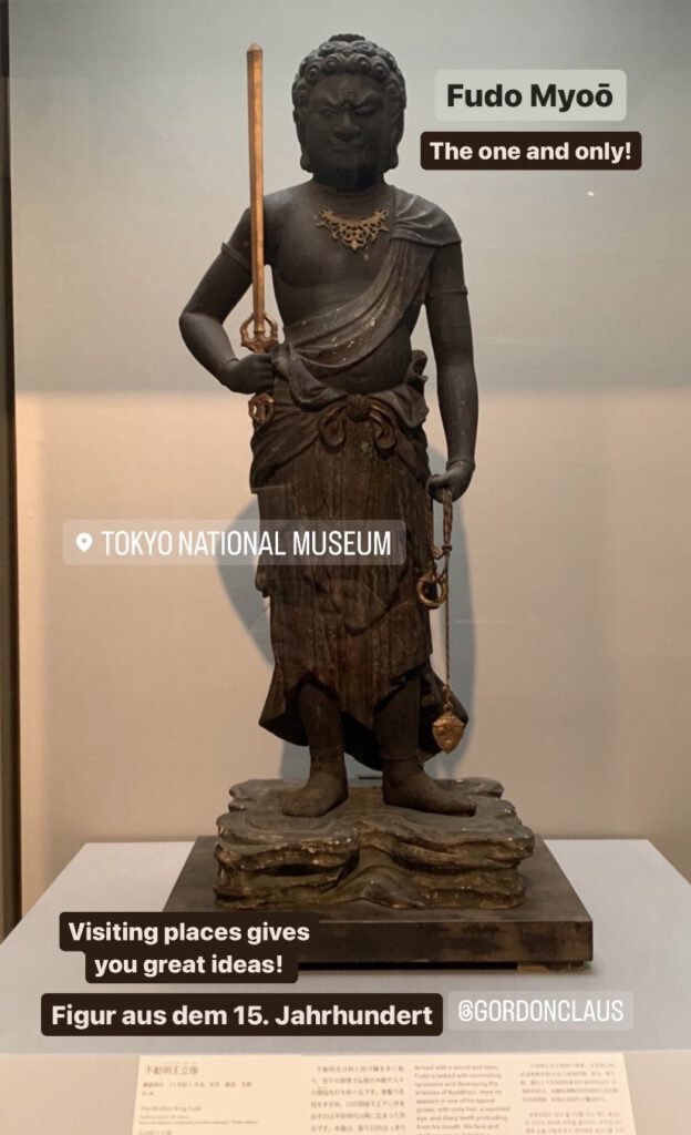 Foto depicting God of fire "Fudo Myoō" standing on rocks holding his sword upright in the right hand. in his other hand he holds a rope. cutting through ignorance and fasting you greed. This picture was taken in the National Gallery Tokio and has the tipical white wall background.