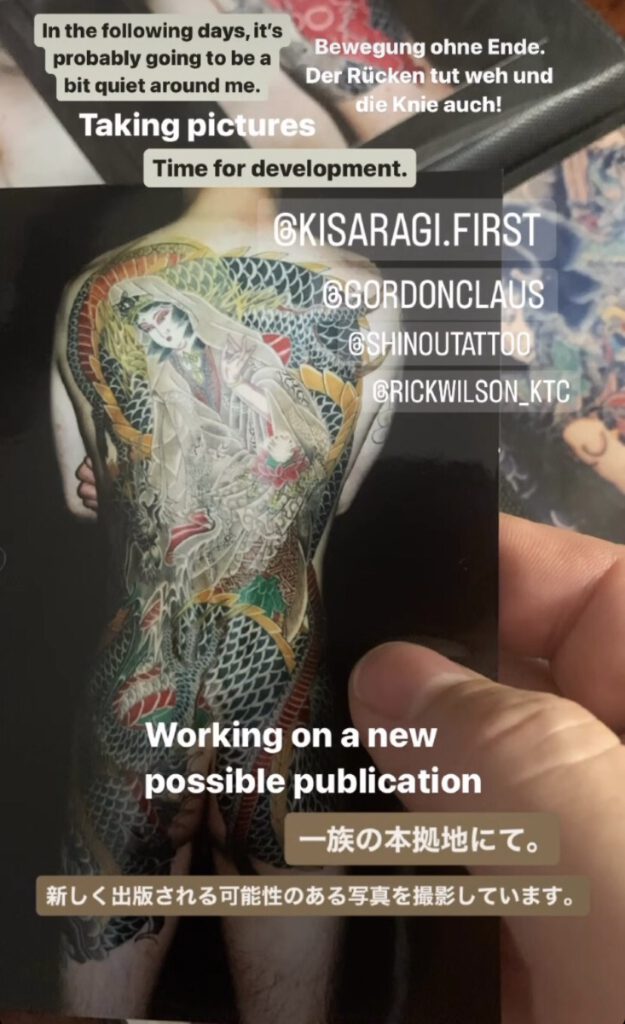 a hand holding a fotograph of a mans back adorned with Senju Kannon, goddess of Mercy and a dragon wrapped around her body. Other fotos laying in the background