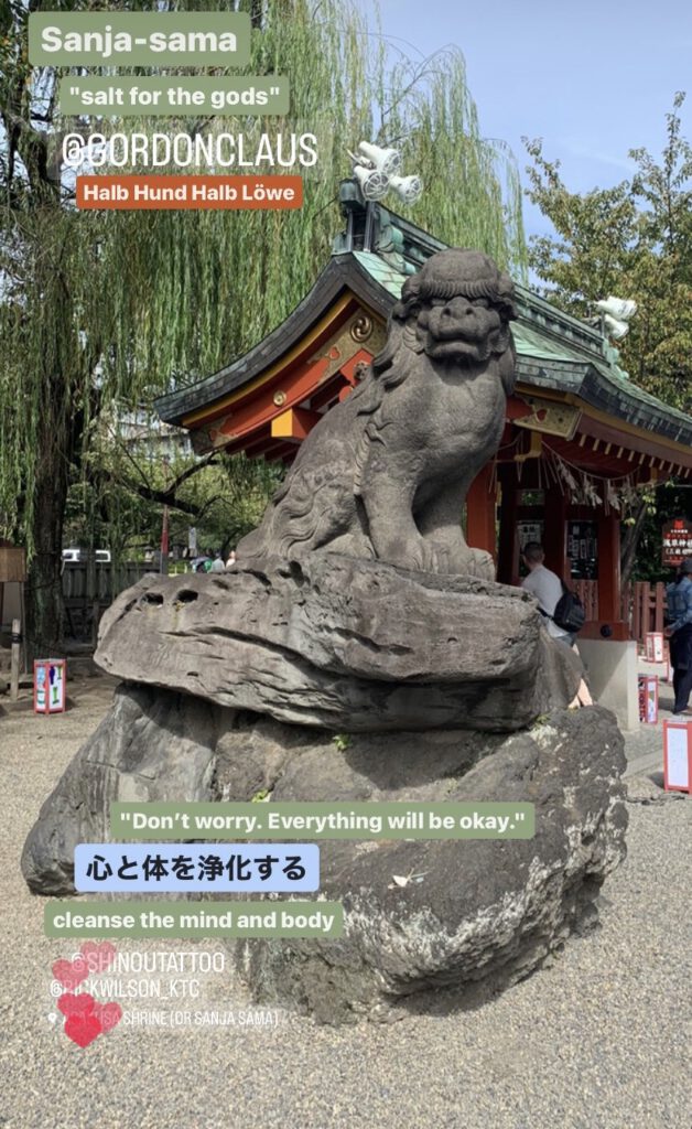 Sanja-Sama "Salt for the dogs" Stone Lion sitting on a pedestal at Asakusa shrine. The statue surrounded by grey pebbles amd a willow tree in the background.