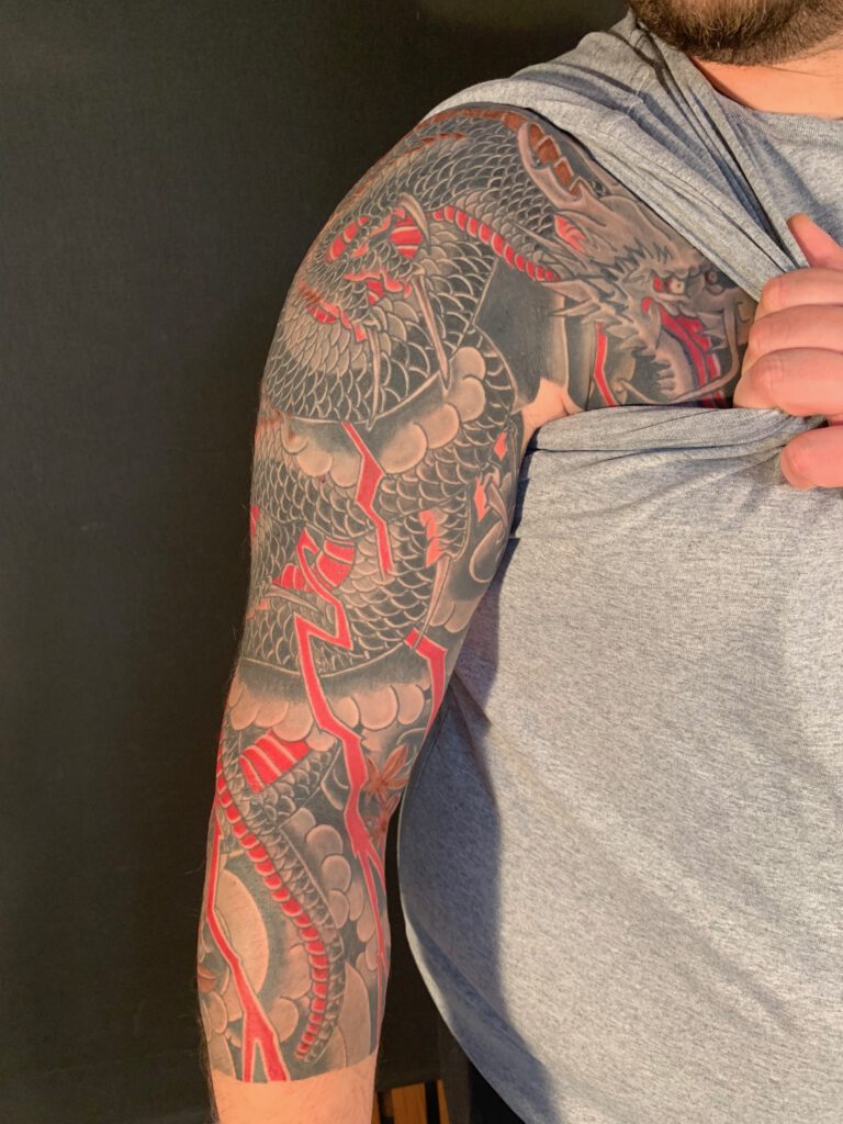 Red and black coiling dragon on mans arm. The man is pulling up this sleeve and the dragons head on the chest becomes visible.