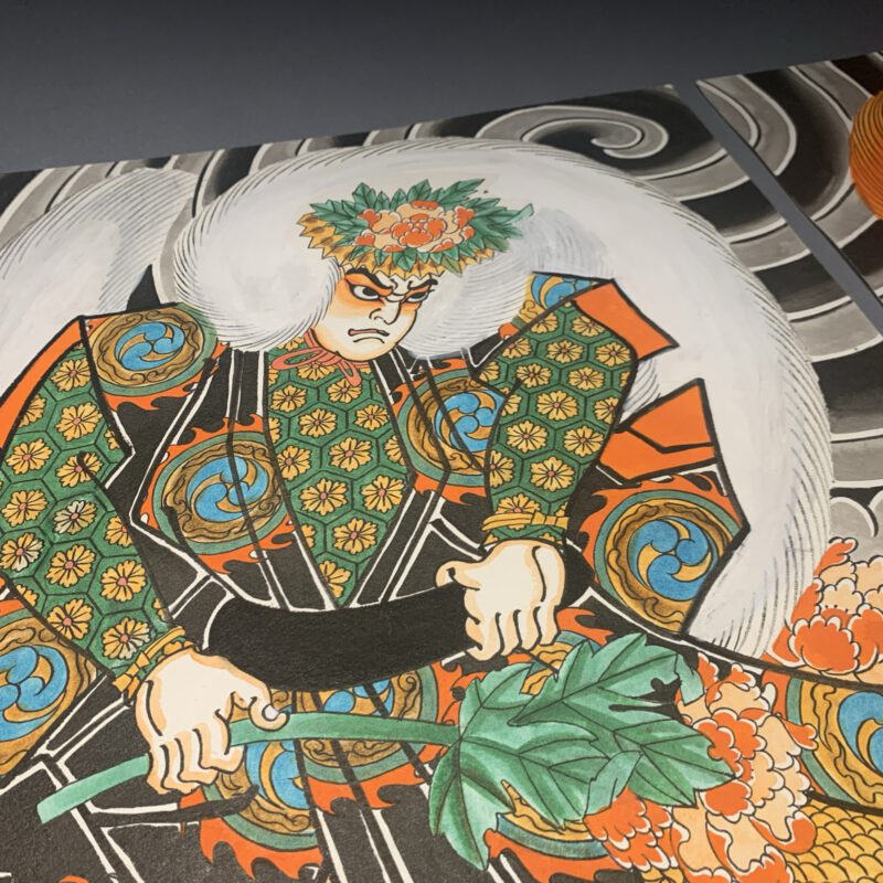 Two Paintings of Kabuki Actors on a gray table. The actors representing Lion dancers from Japanese theater are presented with beautiful garment Design wearing Wings made of Jak ox hair. One has a white wig and the other a red wig.